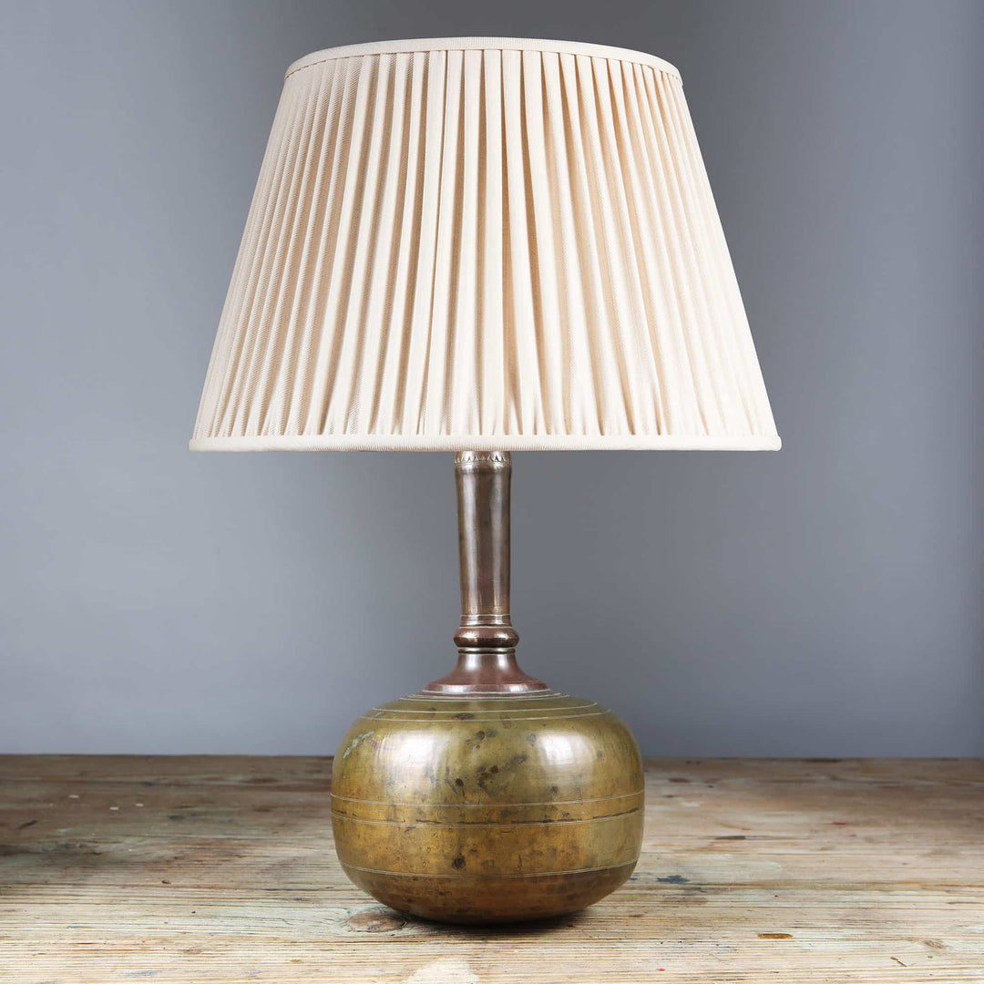Rajasthani brass water bottle as a Table Lamp with a pleated cream lampshade on wooden surface against gray wall