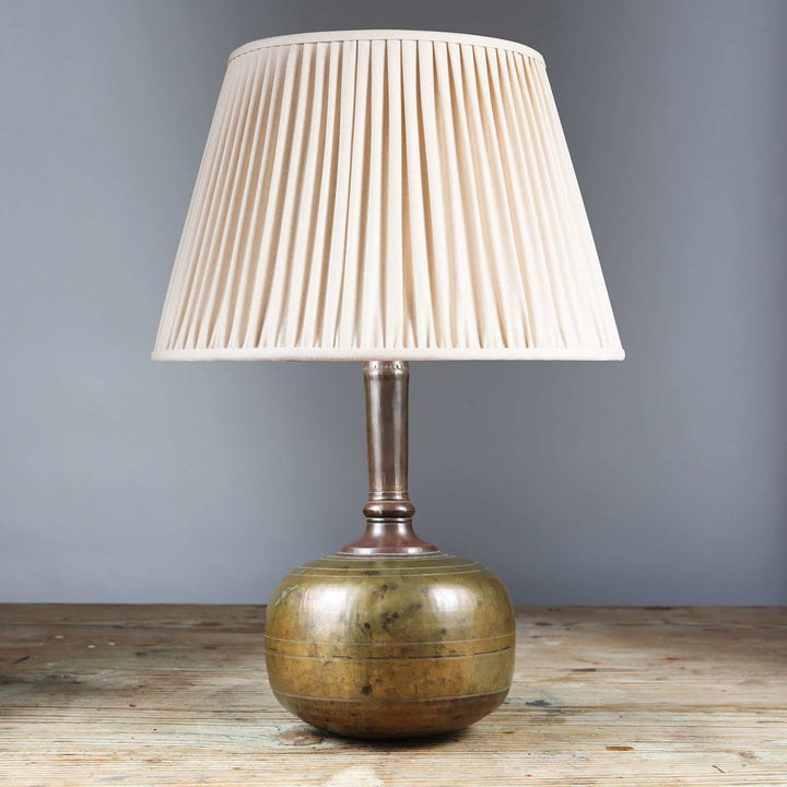 Rajasthani brass water bottle as a Table Lamp with a pleated cream lampshade on wooden surface against gray wall