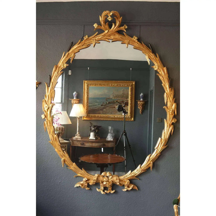 Substantial oval giltwood mirror with elaborate carved frame reflecting an elegant vintage interior setting.