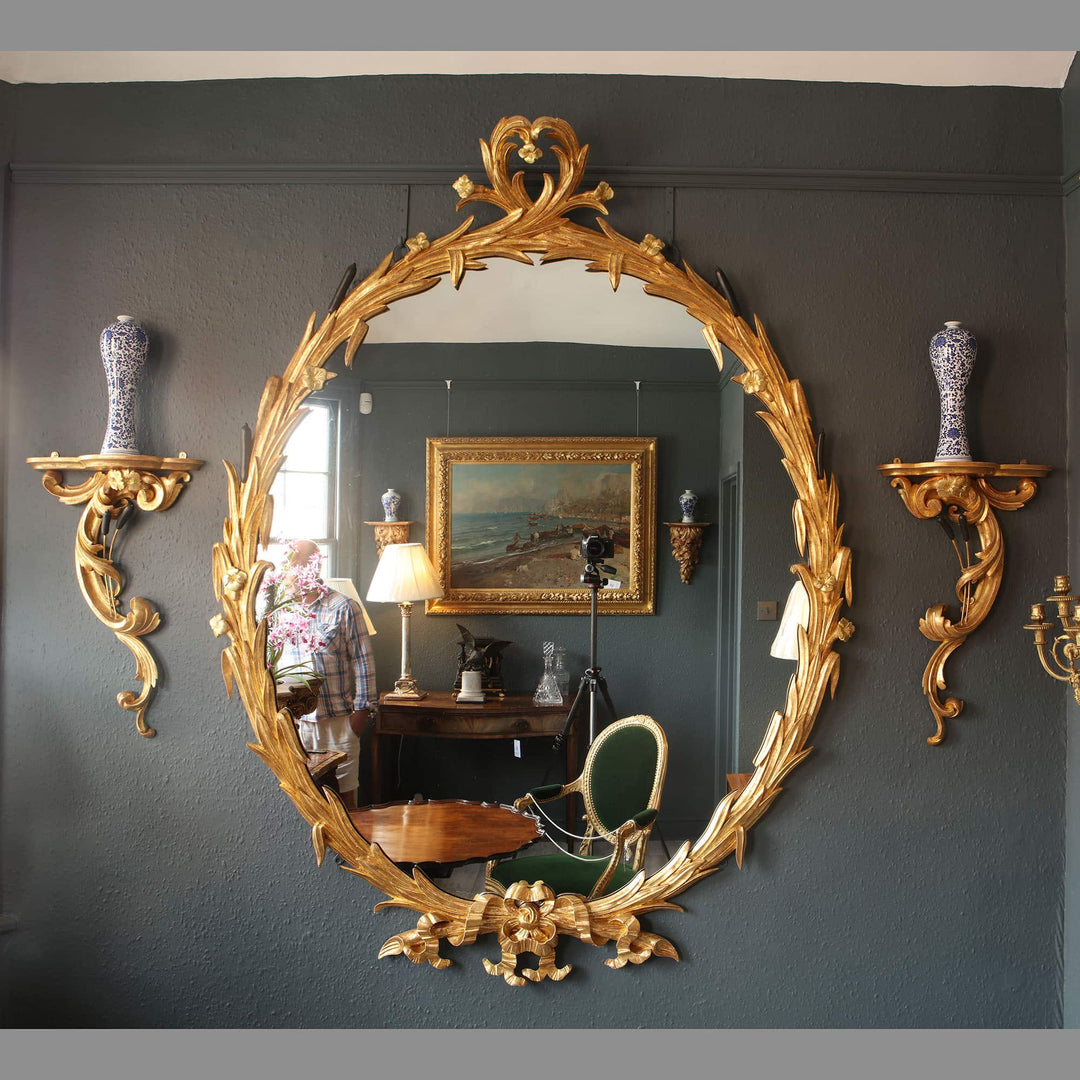 Substantial oval giltwood mirror with ornate gold leaf frame reflecting elegant vintage interior decor.