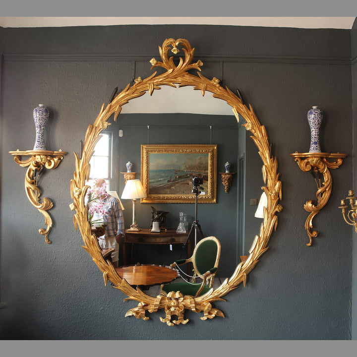 Substantial oval giltwood mirror with ornate gold leaf frame reflecting elegant vintage interior decor.