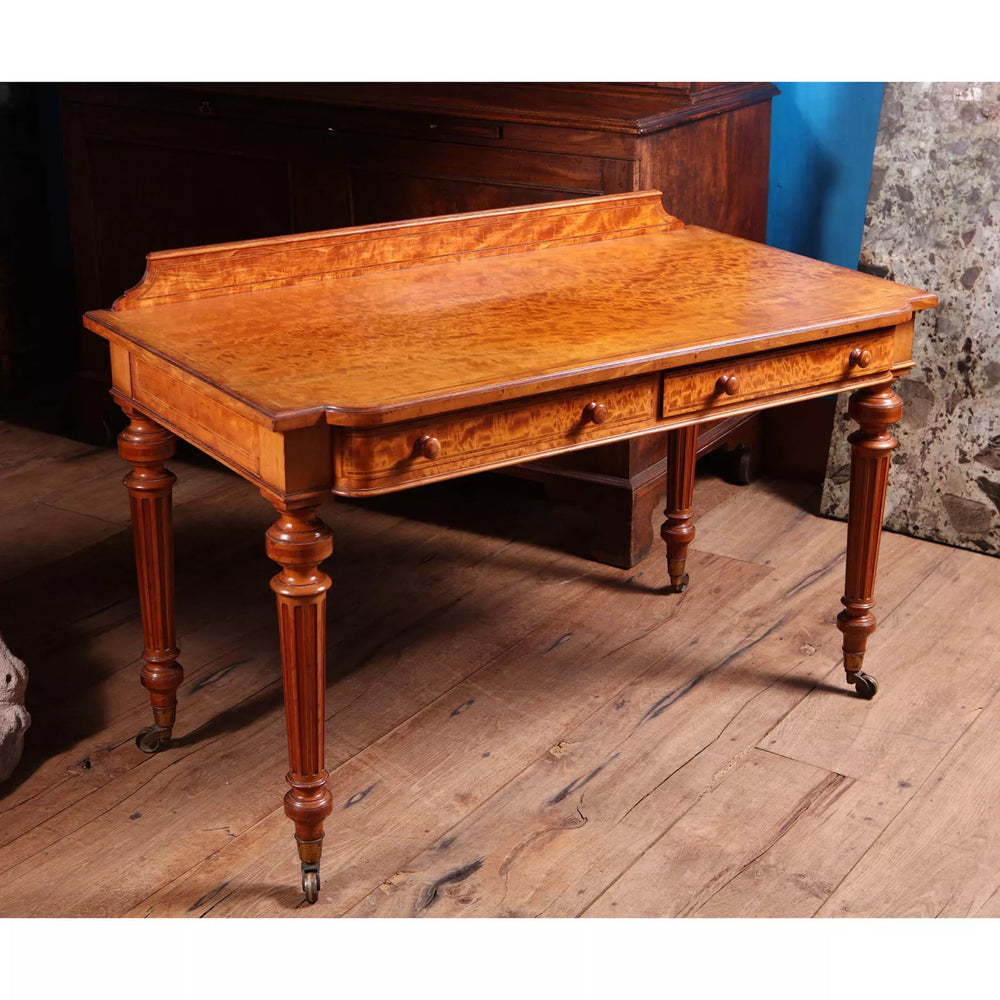 Victorian Satinwood Writing Table by Gillow & Co with figured satinwood top, two drawers, fluted legs, and brass castors.