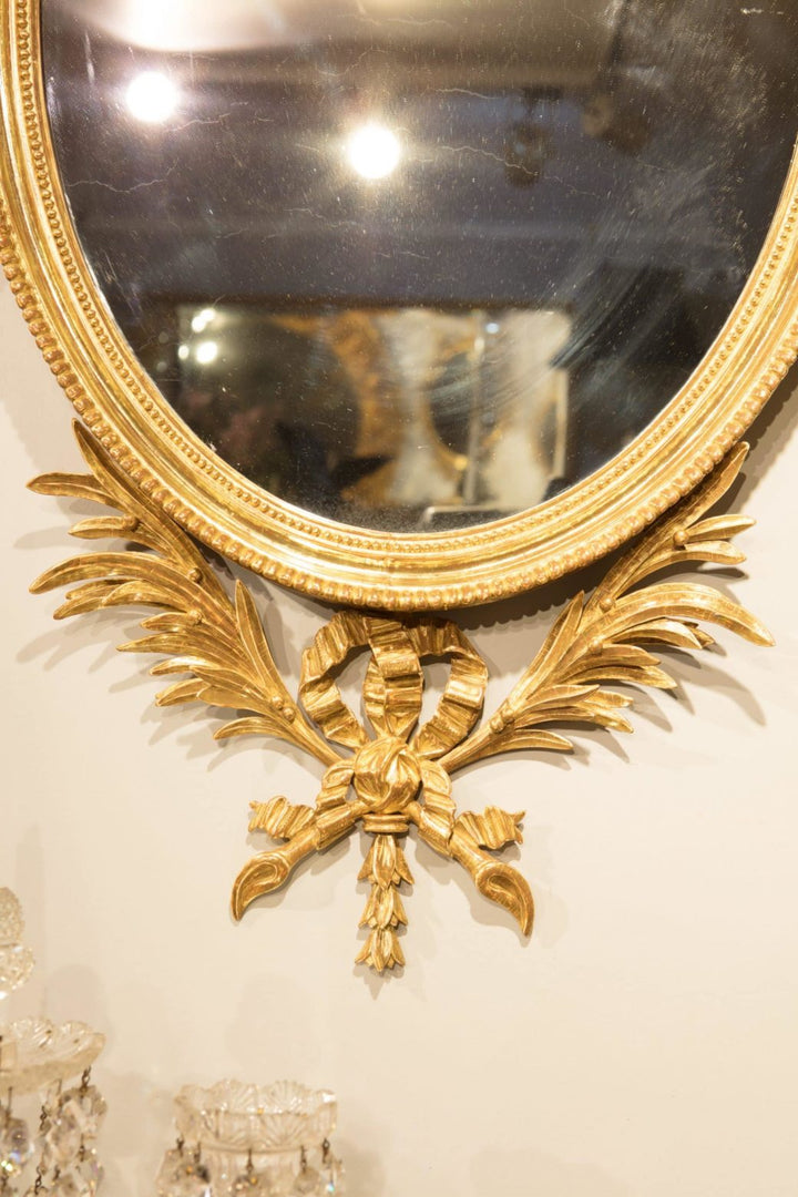 Pair of George III Giltwood Oval Mirrors with ornate palm leaf and bow designs, circa 1770, England, 109 cm high.